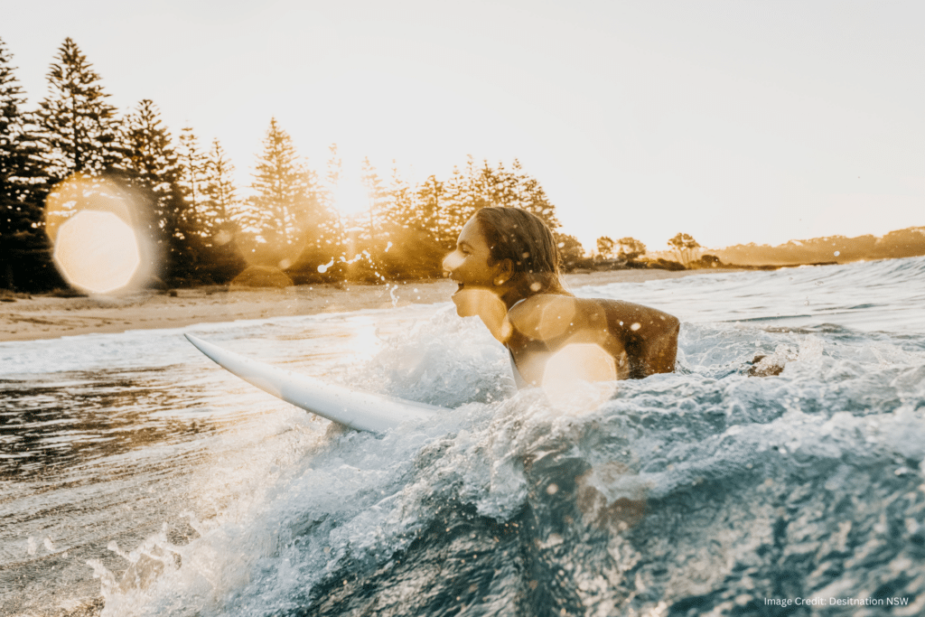 A woman surfing on Durras Beach, with the Joalah Holiday Park campgrounds visible in the background. The image shows the woman riding a wave and enjoying the ocean.